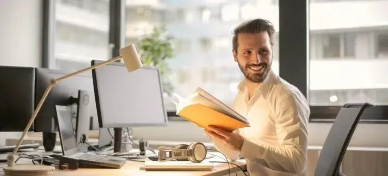 Smiling man holding orange book at office desk