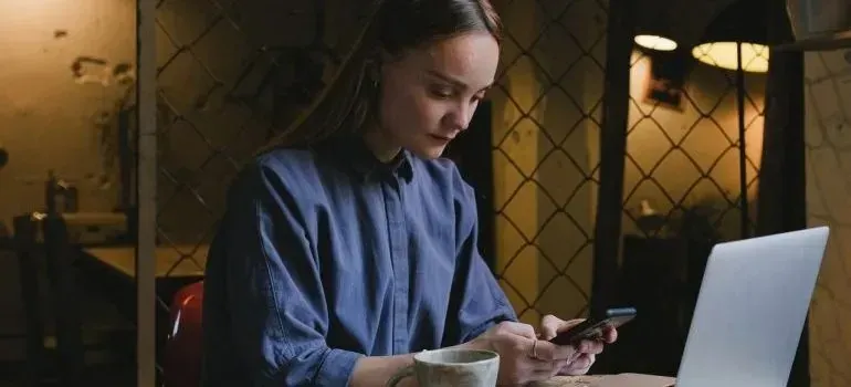 Woman checking phone beside laptop in dim cafe