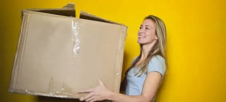 Smiling woman holding large cardboard box, yellow background