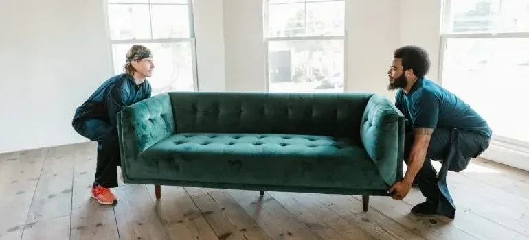 Two movers lifting green velvet sofa in empty room