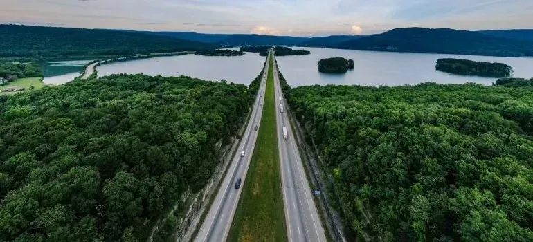 Aerial highway through forest beside lake, distant trucks