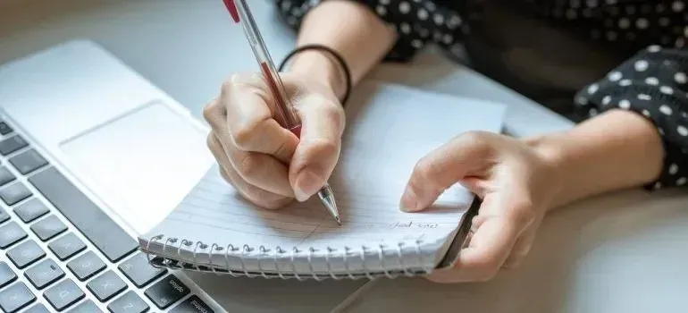 Hand writing checklist in spiral notebook beside laptop