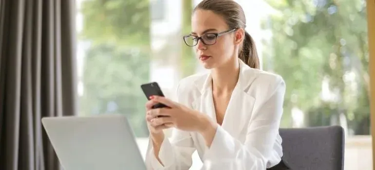 Woman in white blazer using phone beside laptop
