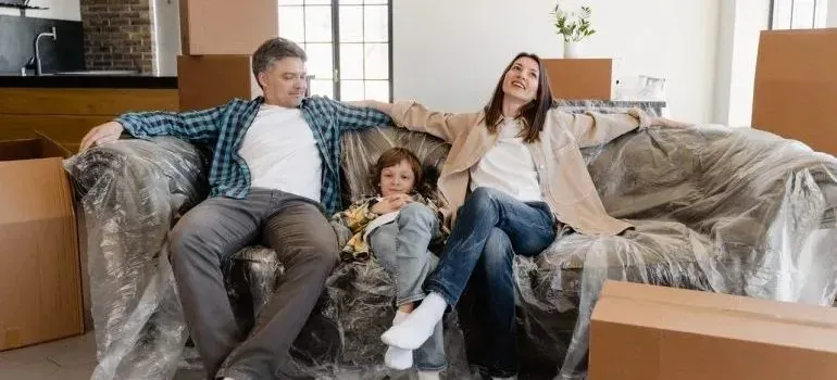 Family resting on plastic-wrapped sofa among moving boxes