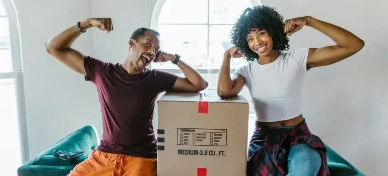 Couple flexing playfully beside medium cardboard moving box