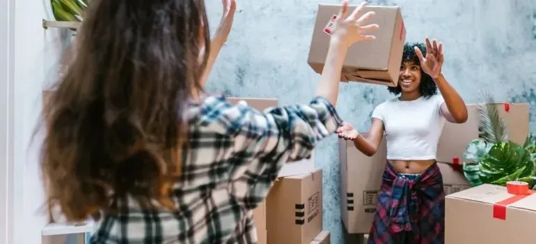Two women playfully tossing cardboard box while unpacking