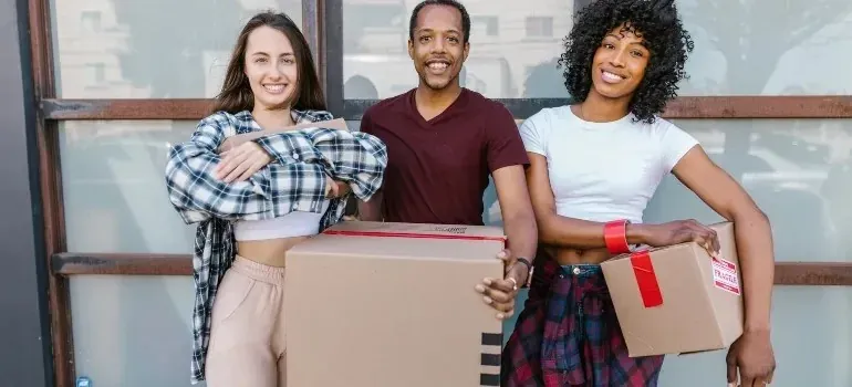 Three smiling friends holding taped moving boxes outdoors
