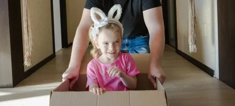Girl in bunny ears riding cardboard box pushed indoors