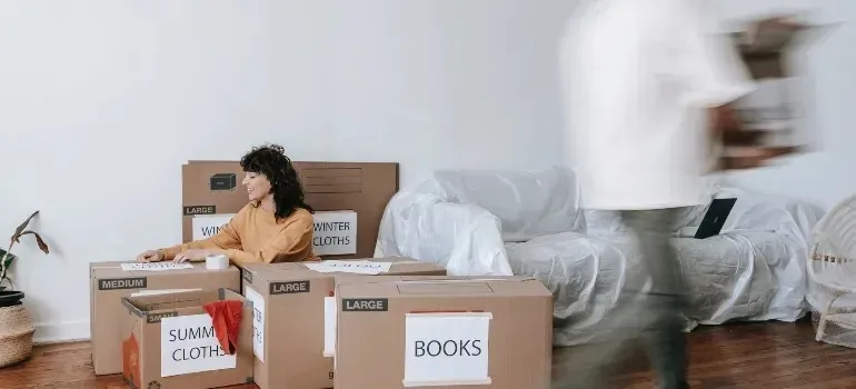 Woman unpacking labeled moving boxes, blurred man walking by