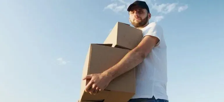 Man in cap carrying two cardboard boxes against sky