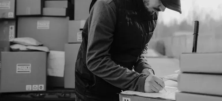 Black-and-white courier writing on clipboard beside stacked boxes