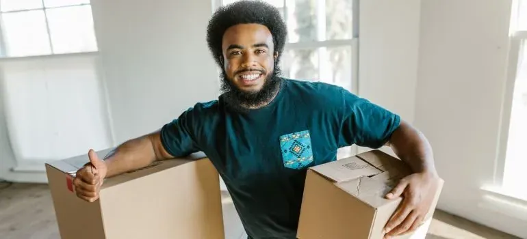 Smiling bearded mover holding two cardboard boxes indoors