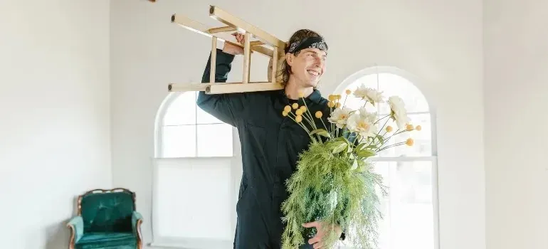 Smiling man carrying wooden stool and flowers indoors