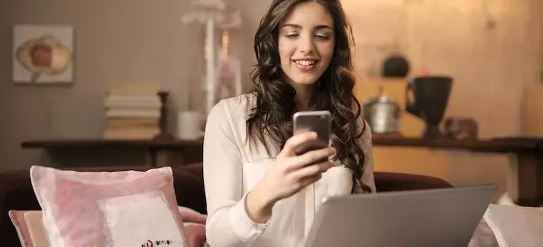 Woman smiling at smartphone with laptop on couch