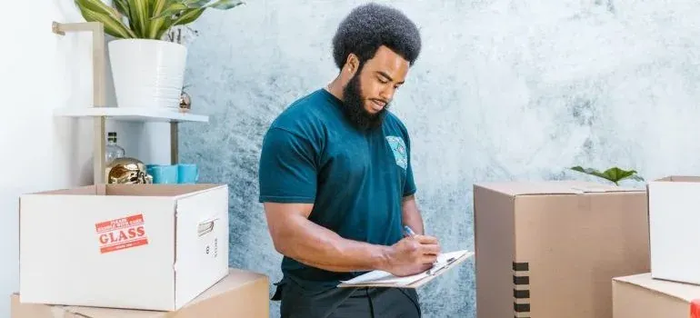 Bearded mover writing inventory on clipboard among boxes