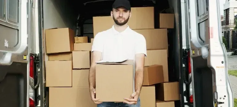 Man in cap holding cardboard box beside loaded cargo van