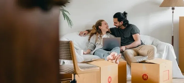 Couple on sofa with laptop surrounded by moving boxes