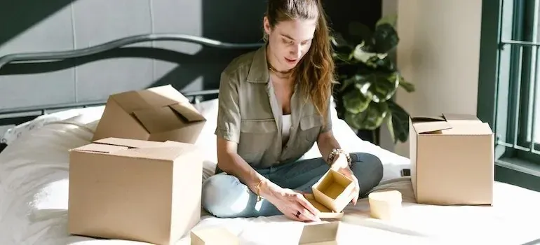 Woman sitting on bed unpacking small boxes in sunlight