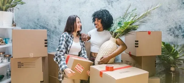 Two women carrying taped moving boxes and a woven vase