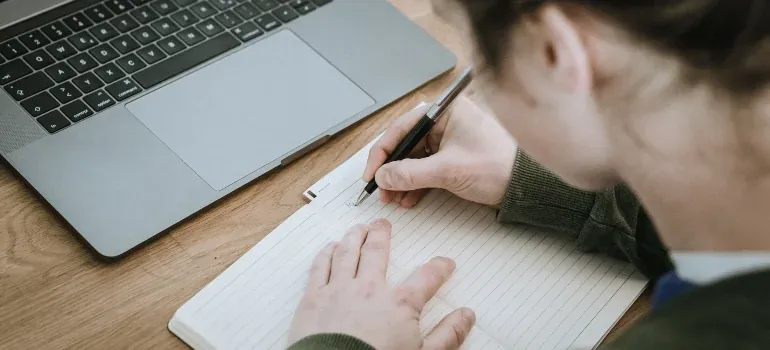 Person writing in notebook by laptop